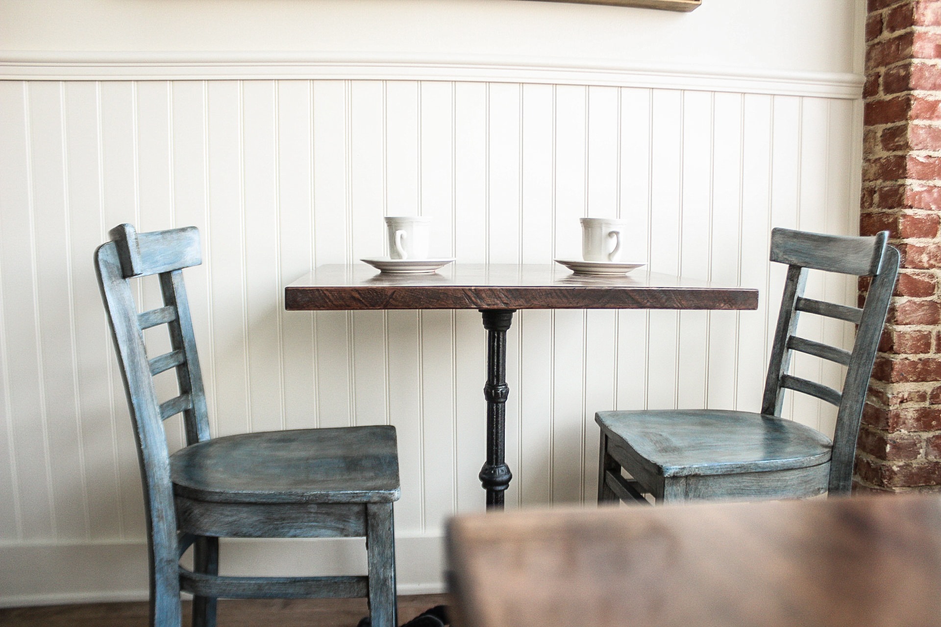 Rustic bistro table with two blue chairs and white paneled wall.